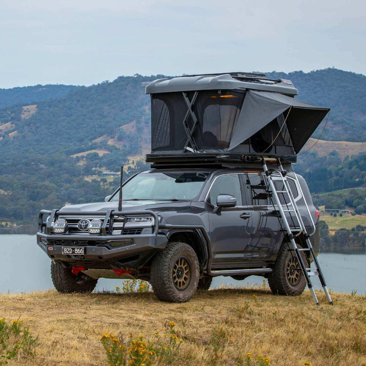 Black SUV with a rooftop tent parked on a grassy area with mountains and water in the background.